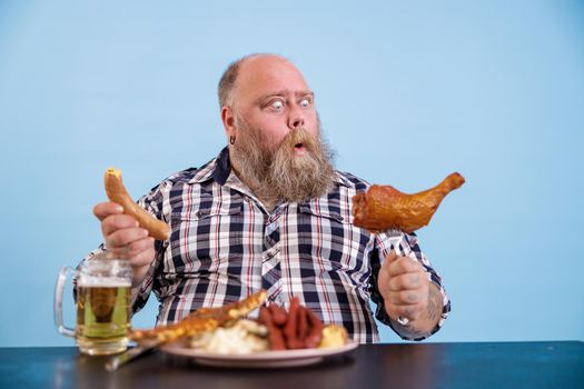 Shocked bearded man looks at chicken leg on fork sitting at table with different fat food and beer on light blue background in studio