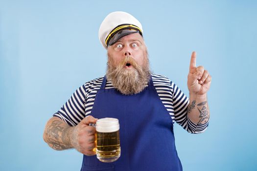 Emotional mature sailor man with overweight in apron and captainhat holds beer and gestures on light blue background in studio