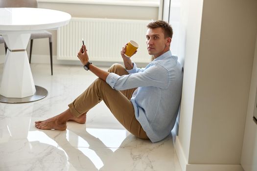 Caucasian hadsome young man in blue shirt is sitting on the floor and chilling at modern kitchen