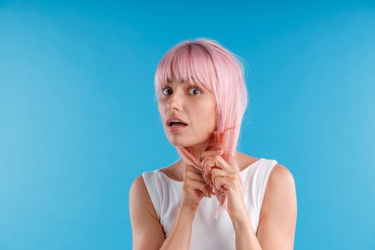 Portrait of surprised female model playing with pink hair, touching it while posing isolated over blue studio background. Beauty, hair care concept