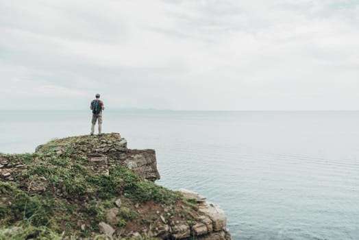 Traveler young man with backpack standing on edge of cliff coast and enjoying view of sea.