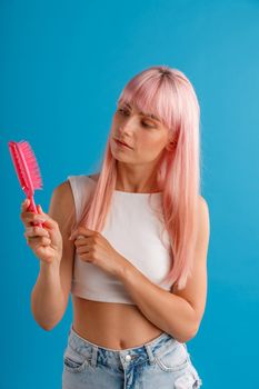 Curious woman with smooth natural long pink hair looking at hair comb while standing isolated over blue studio background. Beauty, hair care concept