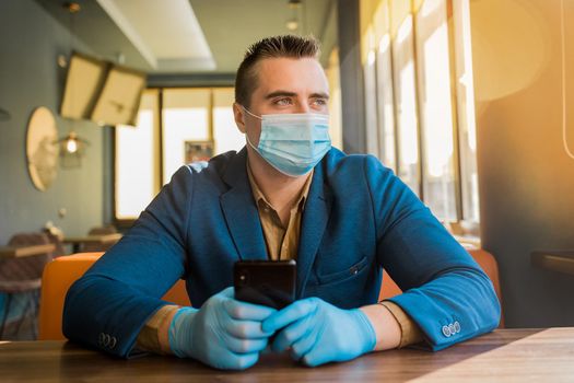 Young european guy businessman in protective gloves and a medical mask spends time on the phone and looks out the window.
