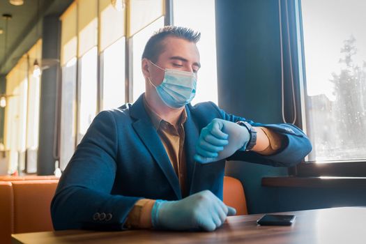 Young attractive guy businessman in medical gloves and a protective mask looks at the time on a wristwatch.