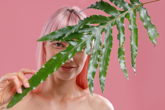 Young woman with pink hair looking at camera, hiding behind green plant leaf, posing isolated over pink studio background. Beauty, skin care, natural cosmetics concept