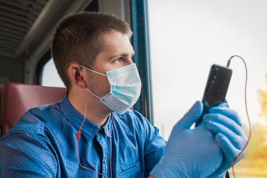 A guy in a protective mask and medical gloves is holding a phone and listening to music on headphones looking out the window.