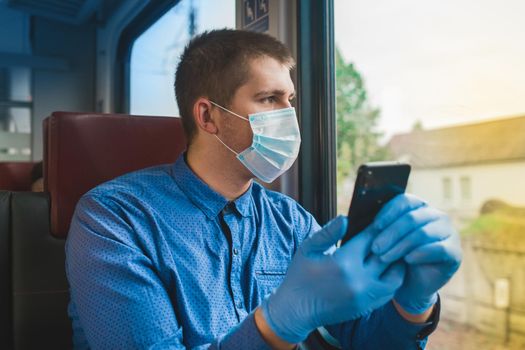 Young european guy in a blue shirt, protective gloves and a medical mask with a phone in his hands looks out the window of a modern electric train.