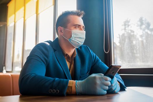 Caucasian male businessman in protective gloves and medical mask spends time in mobile phone in cafe.