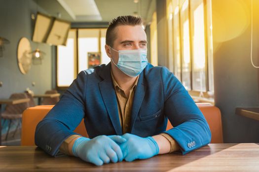 European man businessman in protective gloves and a medical mask sits at a table in a cafe and looks out the window.