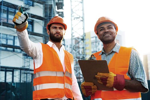 Two young male engineers in uniform and hardhats working at construction site, close up