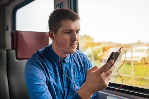 A young European guy in a blue shirt spends time on the phone listening to music on headphones while traveling on a modern electric train.