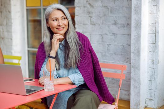 Dreamful senior Asian woman with grey hair sits at coral table with computer and drink on outdoors cafe terrace