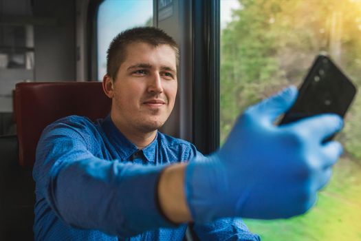 A young guy in a blue shirt and protective medical gloves with a phone in his hands, takes a selfie or talks on a video call in a modern electric train.