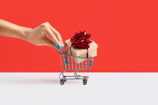 Hand pushing Toy shopping cart or trolley with large gift box. A gift for a holiday, anniversary, or birthday. Trolley cart with a gift box Isolated over red background. Banner with copy space.