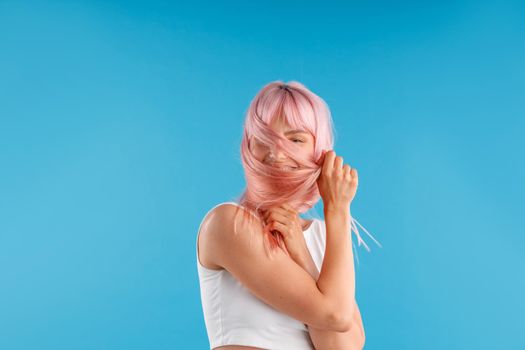 Smiling female model in white shirt playing with smooth straight pink hair, hiding her face while posing isolated over blue studio background. Beauty, hair care concept