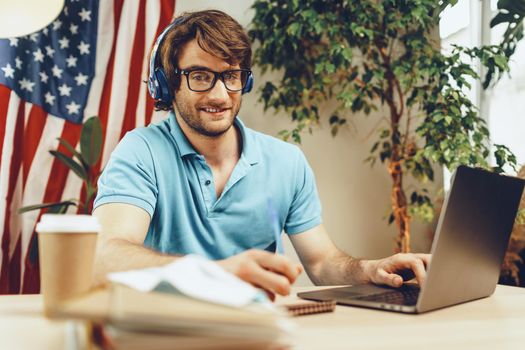 Young bearded businessman sitting at table with laptop and american flag, patriot man