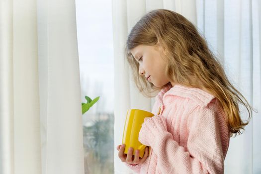 Girl child 7 years old blonde with long wavy hair in a warm knitted sweater holds a cup of tea and looks in the window.