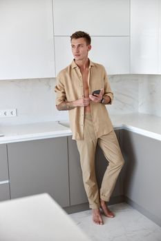 Hadsome smiling man is chilling on the minimalistic kitchen with smartphone