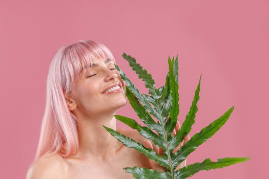Portrait of pleased woman with pink hair smiling with eyes closed, holding plant leaf near her face, posing isolated over pink studio background. Beauty, skin care, natural cosmetics concept