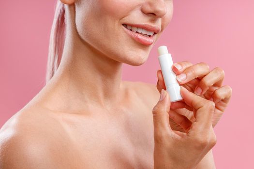 Cropped portrait of smiling woman with naked shoulders holding lip balm near her lips isolated over pink background. Beauty, lip care concept