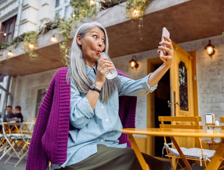 Funny senior Asian woman drinks water through straw taking selfie with cellphone at cafe table outdoors low angle view