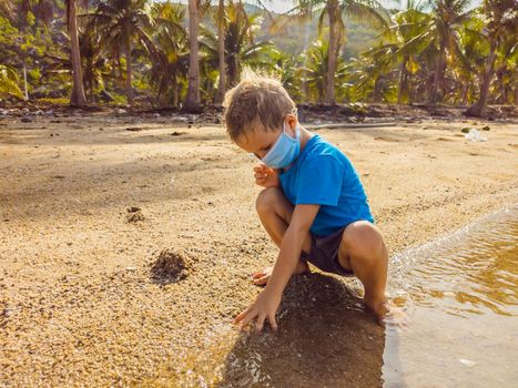 Cute little young blond boy with medical face mask on the beach playing with sand. Protecting from air pollution and virus infection as new normal lifestyle during the covid19 virus epidemic era.
