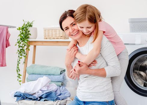 Little girl hugging with mother during laundry of clothes in washing machine at home