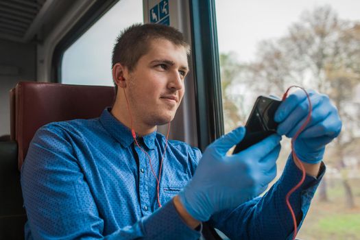 Young european guy in protective medical gloves holds a phone in his hands and listens to music with headphones in a modern electric train.