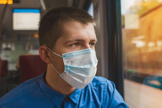 Young Caucasian guy in a protective medical mask looks out the window of a modern electric train at the station.