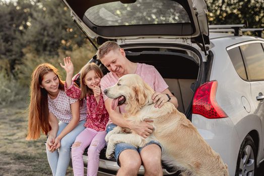 Father with daughters and dog sitting in open car trunk outdoors