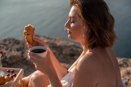 Woman covered with a blanket of bed relaxing and watching the seascape at sunrise. She holds a cup of coffee in her hand in front of her is a table with fruits and croissants. Wanderlust and freedom concept scene