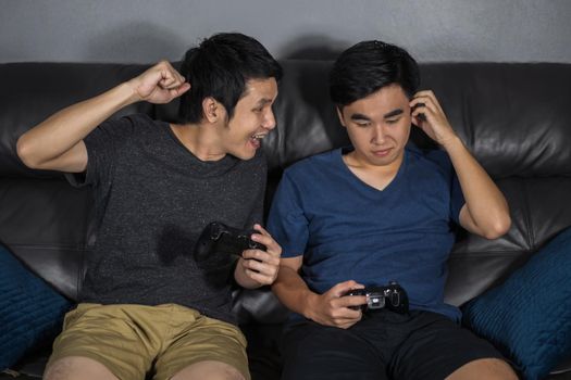two young man playing video games while sitting on sofa. win and lose emotions