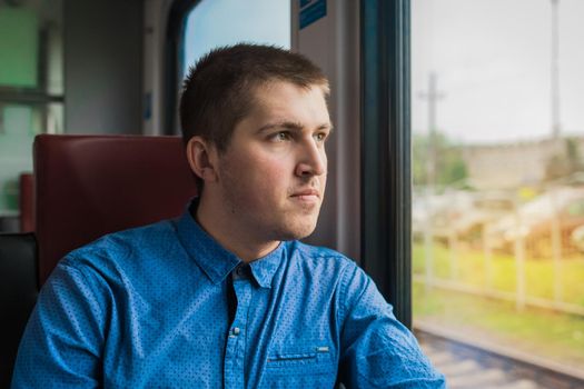 Young european guy in blue shirt looking out the window of a modern electric train traveling.