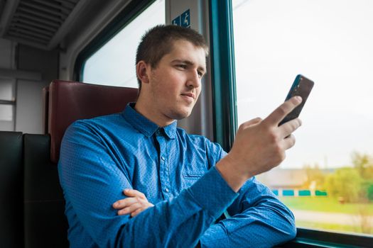 Young european brunette guy in a blue shirt with a grin looks at a mobile phone in his hand in a modern electric train traveling.