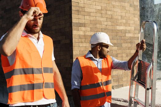 Two young male engineers in uniform and hardhats working at construction site, close up