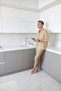 Hadsome smiling man is chilling on the minimalistic kitchen with smartphone