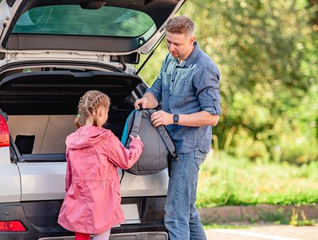 Father giving backpack to school girl next to open car trunk