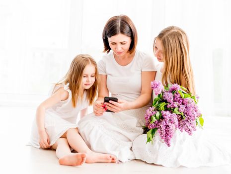 Mother with two little daughters sitting on the floor in the bright room and looking at the smartphone. Beautiful mom with two little daughters in white dresses looking on photos at smartphone