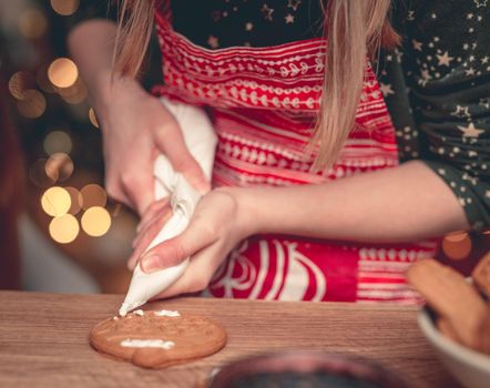 Girl in festive apron decorating gingerbread using culinary bag at home