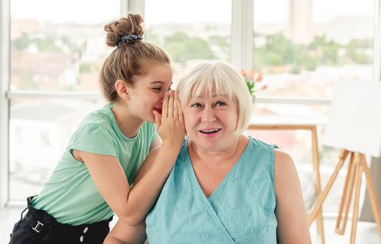 Happy little girl whispering secret to smiling grandmother in bright room