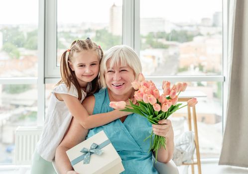 Smiling granny with presents from cute granddaughter in bright room