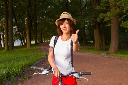 Young woman cyclist in the hat in the park showing thumb up sign