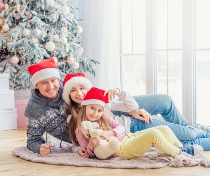 Smiling family in santa hats lying together near christmas tree