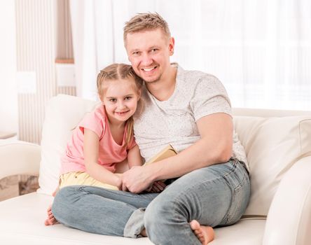 Handsome dad and cutie daughter sitting happy on the beige couch in guest room, with a fathers day gift