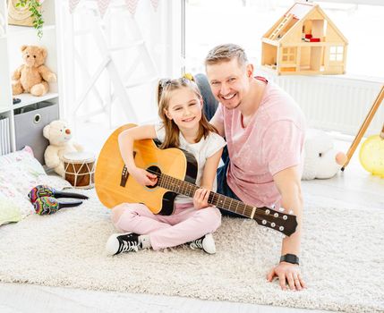 Happy father with cheerful daughter playing guitar in children room