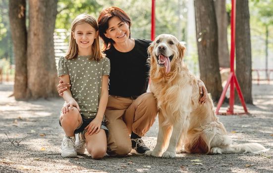 Family with golden retriever dog portrait in the park. Mother, daughter and doggy pet looking at camera together