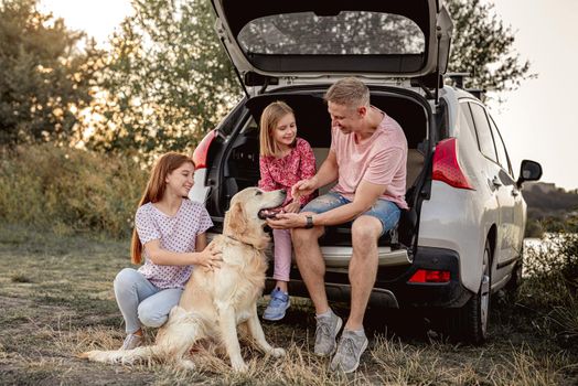 Father with daughters and golden retriever sitting in car trunk on nature