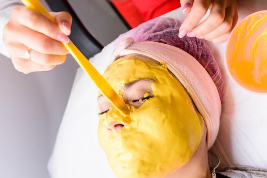 spa treatments in a beauty salon, hand of a professional beautician applying a gold mask on the client's face. new