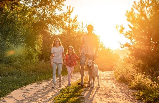 Father with daughters standing holding hands and golden retriever on leash outdoors at sunset