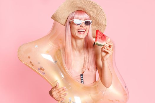 Excited woman with pink hair in swimsuit and sunglasses with watermelon slice and inflatable star for swimming posing isolated over pink background. Summer vacation, fun, party concept